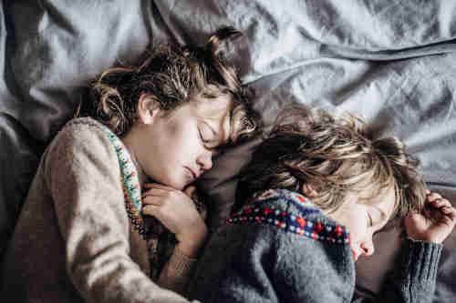 Enfants dormant paisiblement côte à côte Deux enfants endormis l’un à côté de l’autre dans un lit, illustrant l’importance d’un bon oreiller pour le confort et le sommeil des plus petits.